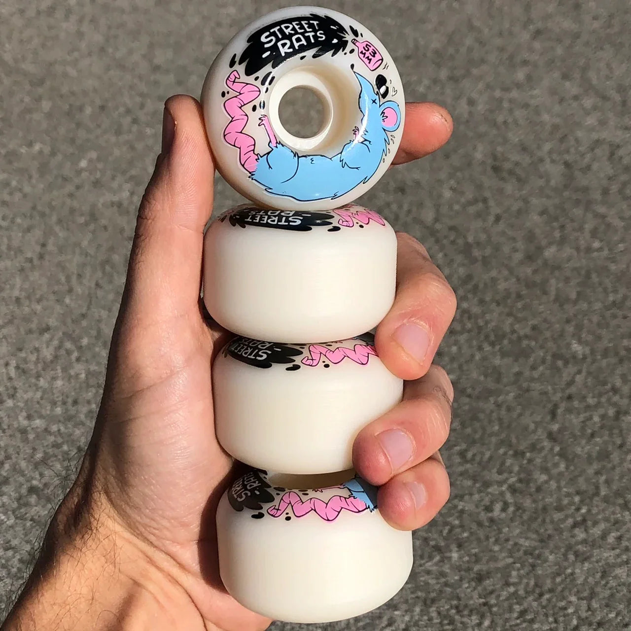 Street Rats Skateboard Wheels; White urethane construction for smooth performance; Features a blue and pink rat with a spilled vial graphic; Provides excellent grip and durability for street skateboarding; Set displayed in hand to show size;